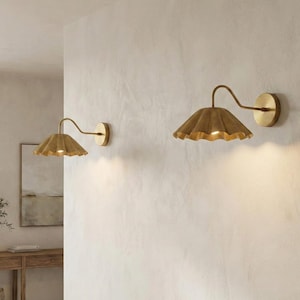 May include: Two wall-mounted sconces with a warm, golden finish. Each sconce features a ruffled, petal-shaped shade and a curved arm extending from a circular wall plate. The lights are turned on, emitting a soft glow.
