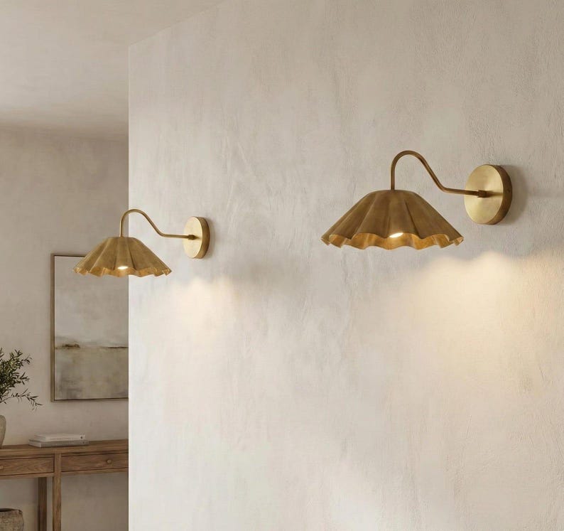 May include: Two wall-mounted sconces with a warm, golden finish. Each sconce features a ruffled, petal-shaped shade and a curved arm extending from a circular wall plate. The lights are turned on, emitting a soft glow.