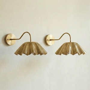 May include: Two brass wall sconces with a scalloped shade design. Each sconce features a circular wall mount, a curved arm, and a ruffled shade. The fixtures are a warm gold color, adding a touch of elegance to any space.