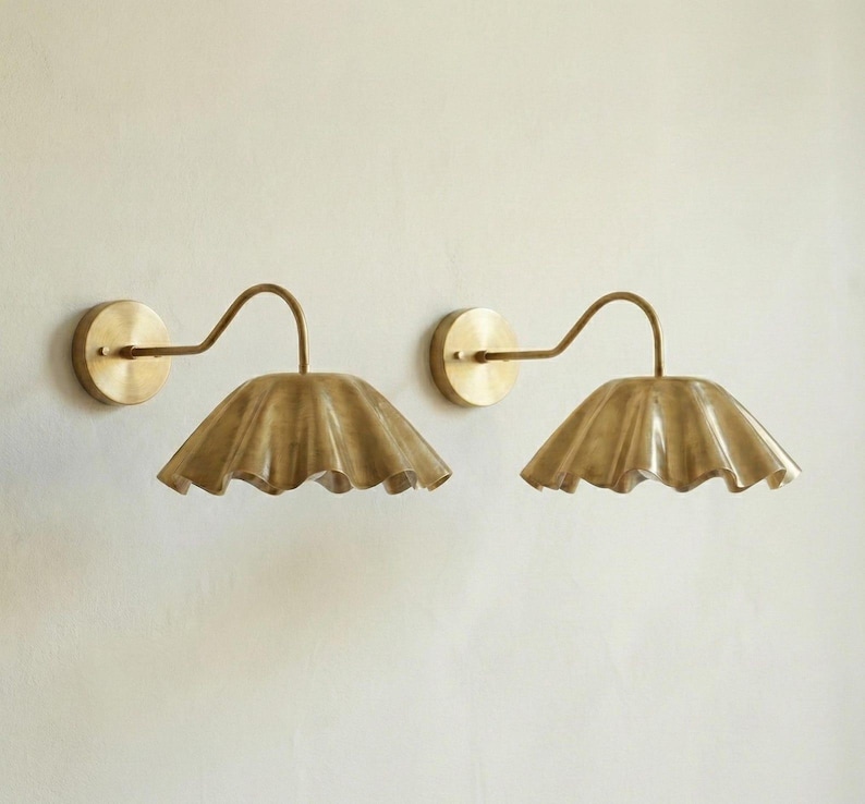 May include: Two brass wall sconces with a scalloped shade design. Each sconce features a circular wall mount, a curved arm, and a ruffled shade. The fixtures are a warm gold color, adding a touch of elegance to any space.