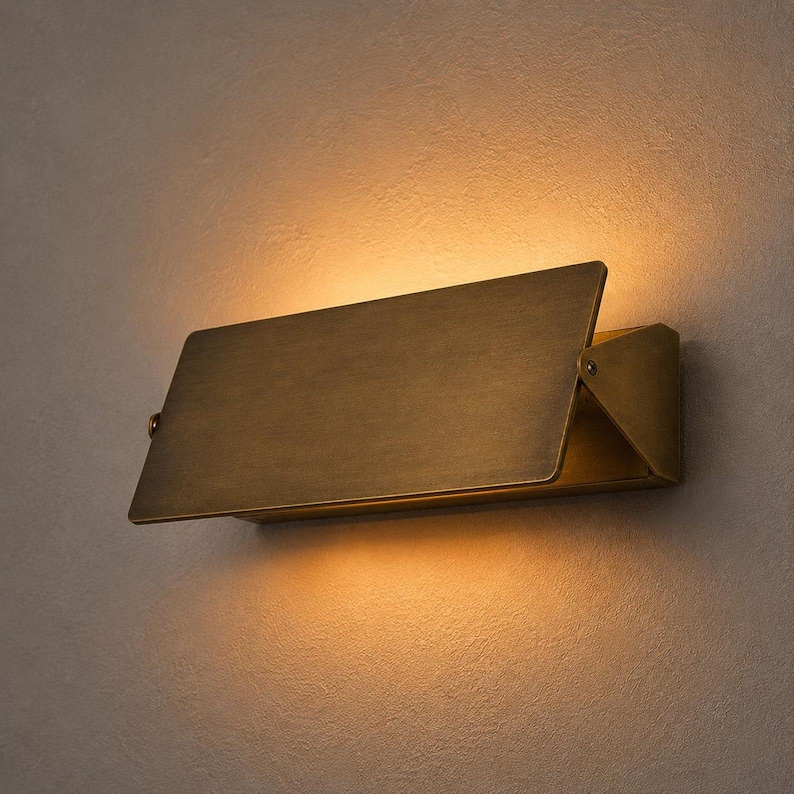 Op de afbeelding: Een rechthoekige wandlamp met een geborstelde messing afwerking. Het armatuur heeft een plat, schuin geplaatst voorpaneel dat warm, gloeiend licht zowel naar boven als naar beneden richt, wat zorgt voor een zachte verlichting op de muur.