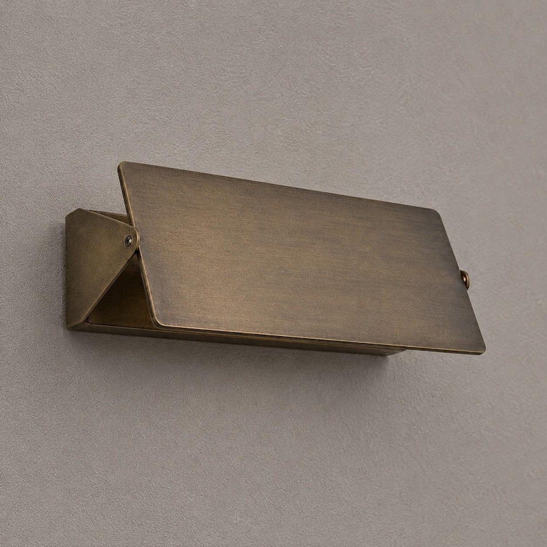 Op de afbeelding: Een rechthoekige wandlamp met een antieke messing afwerking. Het ontwerp heeft een plat, schuin voorpaneel dat uitsteekt vanaf een driehoekige zijbeugel, wat zorgt voor een strak, geometrisch profiel met een metalen, geborstelde textuur.