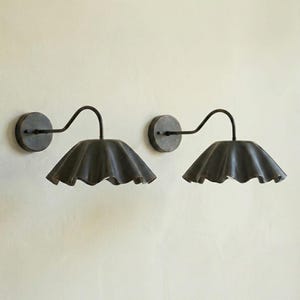 May include: Two dark metal wall sconces with a curved arm and a ruffled shade. The sconces are mounted on a light-colored wall. The shades have a scalloped edge.