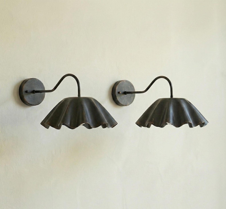 May include: Two dark metal wall sconces with a curved arm and a ruffled shade. The sconces are mounted on a light-colored wall. The shades have a scalloped edge.