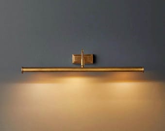 May include: A horizontal wall-mounted picture light fixture with a metallic brass finish. The design features a central rectangular mounting plate and a long, slender cylindrical tube that emits a warm, downward-facing glow across the wall.