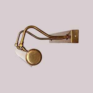 May include: A brass-toned wall-mounted light fixture with a curved arm design. The fixture features a cylindrical shade and a rectangular base. The light is designed to be mounted on a wall and provides focused illumination.