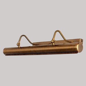 May include: A vintage-style picture light with a bronze finish. The light has a cylindrical body with two curved arms and a rectangular mounting plate. The fixture is designed to illuminate artwork.