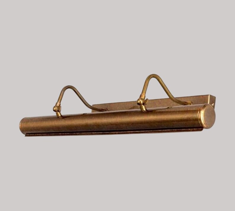 May include: A vintage-style picture light with a bronze finish. The light has a cylindrical body with two curved arms and a rectangular mounting plate. The fixture is designed to illuminate artwork.