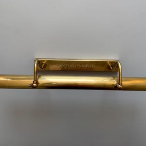 Puede incluir: Una lámpara de latón para cuadros con una placa de montaje rectangular y una carcasa de luz cilíndrica. La lámpara tiene un tono dorado cálido y está montada en una pared gris. El texto "UKFROSTLIGHTSHOPLTD" está grabado.