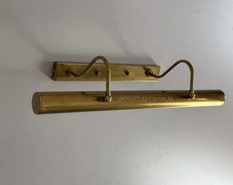 May include: A brass-toned picture light fixture with a rectangular backplate and a curved arm supporting a cylindrical light. The fixture is mounted on a white wall. The text "UKFROSTLIGHTSHOPLTD" is visible.
