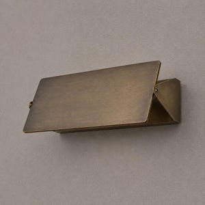 Op de afbeelding: Een rechthoekige wandlamp met een geborstelde antiek messing afwerking. Het ontwerp heeft een plat, schuin voorpaneel dat uitsteekt vanaf een driehoekige zijbeugel, wat zorgt voor een strak, geometrisch profiel met zichtbare metalen schroeven.