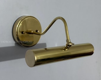 May include: A brass-colored picture light fixture mounted on a wall. The fixture has a circular base, a curved arm, and a cylindrical light housing. The text "UKFROSTLIGHTSHOPLTD" is printed on the base.