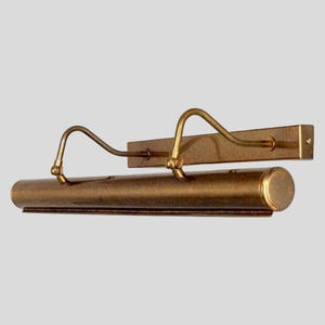 May include: A vintage-style picture light fixture with a warm brass finish. The fixture features a rectangular backplate, a cylindrical light housing, and two curved arms. The design is simple and elegant, suitable for illuminating artwork or photographs.