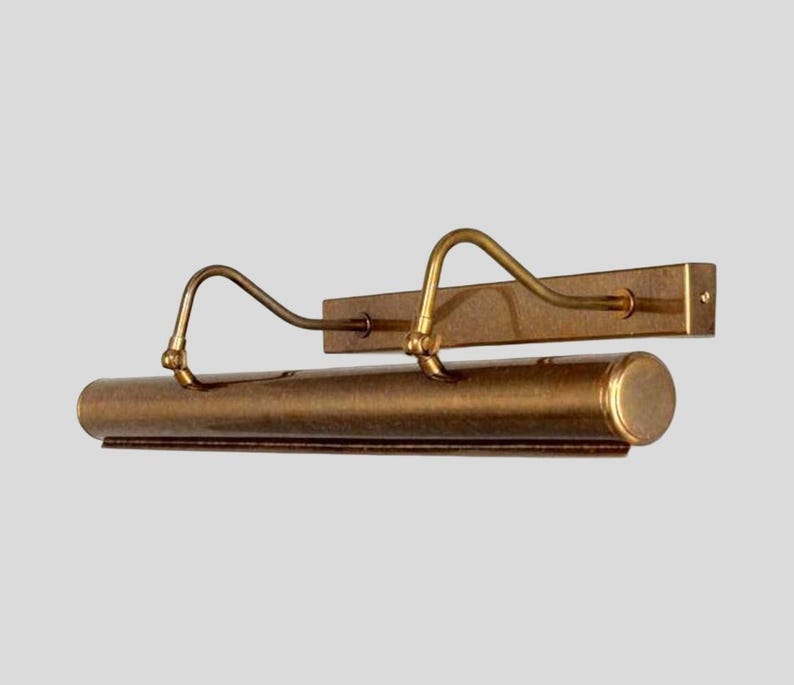 May include: A vintage-style picture light fixture with a warm brass finish. The fixture features a rectangular backplate, a cylindrical light housing, and two curved arms. The design is simple and elegant, suitable for illuminating artwork or photographs.