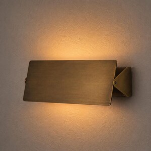 Op de afbeelding: Een rechthoekige wandlamp met een geborstelde messing afwerking. Het armatuur heeft een platte, horizontale metalen plaat bevestigd aan een driehoekige zijbeugel, die een warme, zachte gloed uitstraalt vanaf de boven- en onderkant van de plaat.