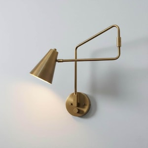 May include: A brass wall sconce featuring a conical lampshade and an adjustable arm. The sconce has a circular base and a geometric arm design. The fixture is mounted on a white wall, providing focused lighting.