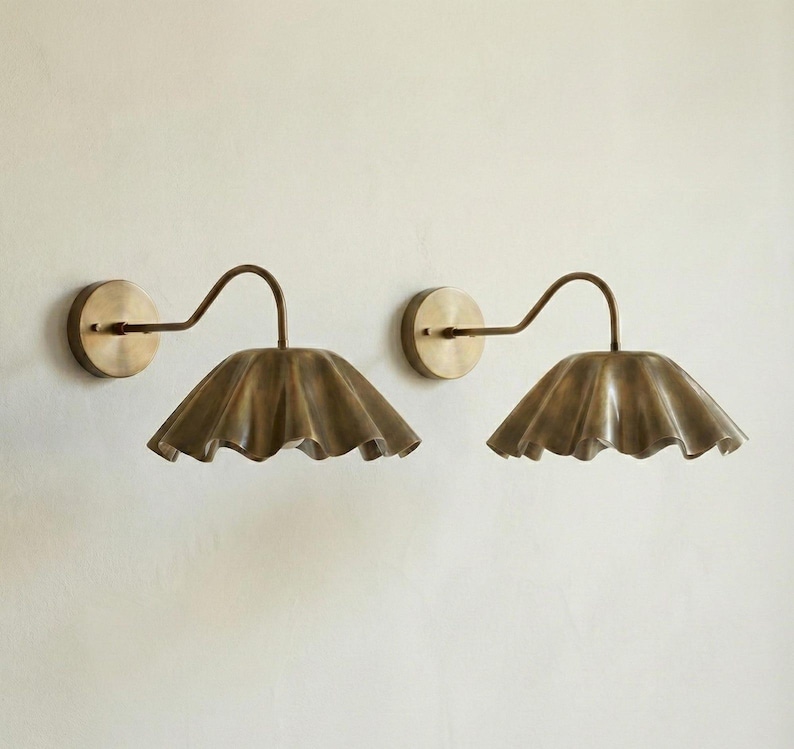 May include: Two wall sconces with a vintage brass finish. Each sconce features a circular wall mount, a curved arm, and a ruffled, scalloped shade. The fixtures are mounted on a light-colored wall, providing a warm, ambient light.