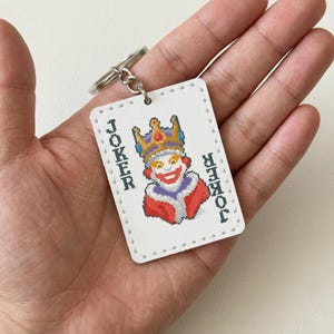 May include: White rectangular keychain featuring a pixelated Joker card design. The Joker wears a crown and red and purple attire. The word "JOKER" is printed vertically on both sides of the card. A silver keyring is attached.
