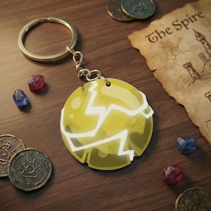 May include: A yellow and white keychain with a cracked design, attached to a gold ring. The keychain is surrounded by coins, gemstones, and a map with the text "The Spire".