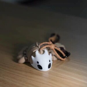 May include: A detailed figurine of a character from the video game Hollow Knight. The figure is primarily beige and white, with black accents on the face. It has a stylized, insect-like appearance and is lying on a wooden surface.