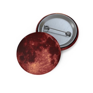 May include: A circular pin-back button featuring a high-resolution photographic print of a blood moon. The surface displays deep crimson and dark orange hues with detailed lunar crater textures. The back shows a silver metal pin mechanism.