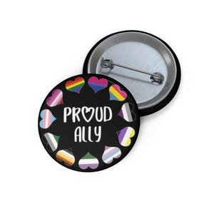 Puede incluir: Un botón redondo con fondo negro que presenta el texto blanco PROUD ALLY en el centro. El texto está rodeado por una disposición circular de once corazones pequeños, cada uno relleno con diferentes patrones de colores de banderas del orgullo.
