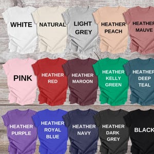 K&ouml;nnte beinhalten: Eine Sammlung von f&uuml;nfzehn kurz&auml;rmeligen T-Shirts mit Rundhalsausschnitt, angeordnet in drei Reihen, jeweils beschriftet mit der Farbe: White, Natural, Light Grey, Heather Peach, Heather Mauve, Pink, Heather Red, Heather Maroon, Heather Kelly Green, Heather Deep Teal, Heather Purple, Heather Royal Blue, Heather Navy, Heather Dark Grey und Black.