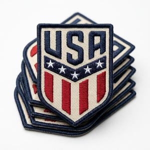 May include: A stack of embroidered patches with a shield design. The patches feature the letters "USA" in navy blue, white stars, and red and white stripes. The patches have a navy blue border and a tan background.
