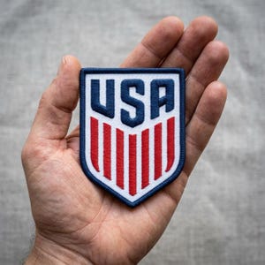 May include: A blue, white, and red USA patch in the shape of a shield. The patch features the letters "USA" in blue at the top, with red and white vertical stripes below. The patch is held in a hand.