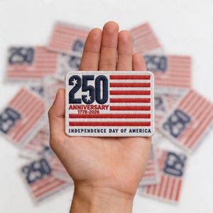 May include: A rectangular patch with a red and white striped design, featuring the text "250 ANNIVERSARY 1776-2026 INDEPENDENCE DAY OF AMERICA" in navy blue and red. The patch is held in a hand, with several similar patches in the background.