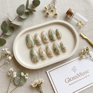 May include: A set of ten almond-shaped press-on nails featuring a sage green and cream marbled pattern with delicate hand-painted white floral and gold leaf accents, displayed on an oval cream tray next to a small glass bottle and a gold tool.