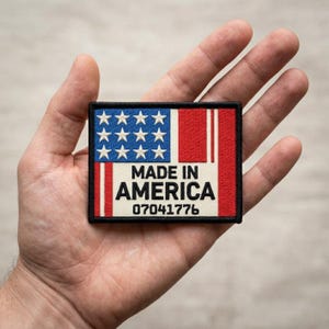 May include: A rectangular patch featuring the American flag design. The upper left corner displays blue with white stars, while the right side has red stripes. The text "MADE IN AMERICA" is centered, with the number "07041776" below.