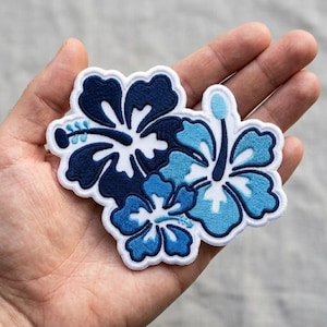 May include: An embroidered patch featuring three hibiscus flowers in varying shades of navy, royal, and light blue. The design includes white outlines and central stamen details, with a white border surrounding the entire floral cluster shape.