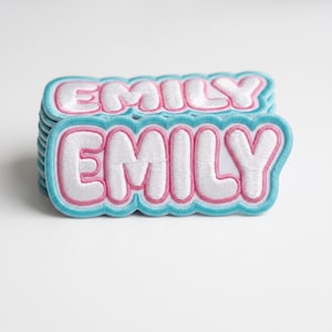 May include: A stack of embroidered patches featuring the name EMILY in white block letters. Each patch has a pink outline and a light blue scalloped border. The design is repeated on every patch in the stack, showcasing the same font and colors.
