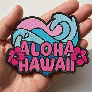May include: A heart-shaped embroidered patch featuring a light blue ocean wave design, the pink text ALOHA HAWAII, and two bright pink hibiscus flowers on the sides, all outlined with a thick black border against a light pink background.