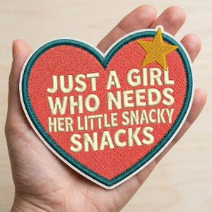May include: A heart-shaped embroidered patch with a red background and a dark teal border. The text reads JUST A GIRL WHO NEEDS HER LITTLE SNACKY SNACKS in cream-colored letters. A yellow five-pointed star is positioned in the upper right corner.