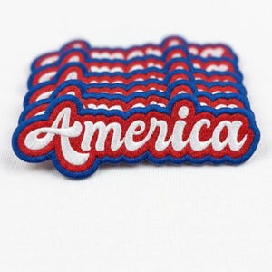 May include: Embroidered patches with the word "America" in white script, outlined in red and blue. The patches are stacked on a white surface, showcasing the patriotic design.