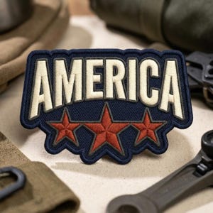 May include: A navy blue embroidered patch with the word "AMERICA" in white block letters. Below the text are three red stars. The patch has a dark blue border and is likely designed to be sewn or ironed onto clothing or gear.