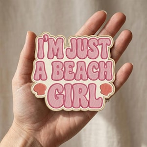 May include: A die-cut embroidered patch with a cream-colored background and pink, wavy-style lettering that reads I'M JUST A BEACH GIRL. The patch features two small, coral-colored seashell icons on the bottom left and right sides of the text.