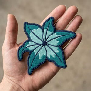 May include: A star-shaped embroidered patch featuring a layered floral design in shades of teal, dark blue, and light mint green. The patch has a dark blue stitched border and a dark blue circular center, with distinct petal-like segments.