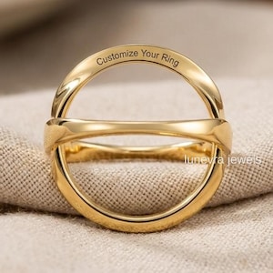 May include: A polished gold-tone ring featuring a circular design with a horizontal bar across the center. The top arc of the ring is engraved with the text "Customize Your Ring" and the lower right side features the text "lunevra jewels".