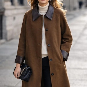 May include: A long brown coat with a leather collar and cuffs. The coat is buttoned up and worn over a cream-colored turtleneck sweater. A black handbag with a chain strap is held in the hand.