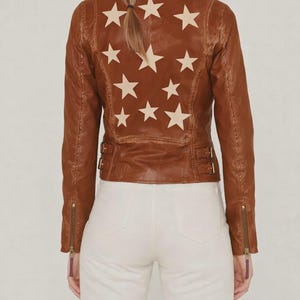Könnte beinhalten: Braune Lederjacke mit Sternmuster auf dem Rücken. Die Jacke hat einen klassischen Biker-Stil mit Reverskragen, Reißverschlussdetails und einer figurbetonten Silhouette. Die Sterne sind cremefarben.