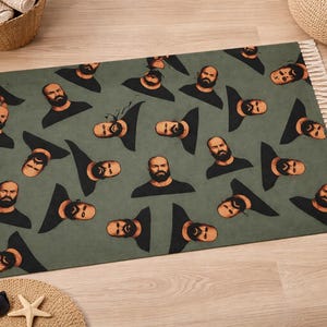 May include: A rectangular rug with a repeating pattern of a man's face and upper torso on a green background. The man has a beard and is wearing a black shirt. The rug has a fringed edge.
