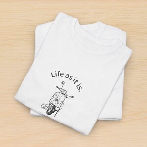 May include: White t-shirts with a graphic of a scooter and the phrase "Life as it is." The design is in black ink. The shirts are folded and stacked on a light wood surface.