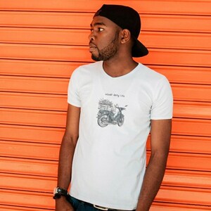 May include: White t-shirt with a graphic of a scooter carrying flowers, and the text "usual daily life". The shirt is worn with a black baseball cap and blue jeans. The background is an orange, horizontally-lined surface.