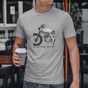 May include: A gray t-shirt featuring a black and white motorcycle illustration. The text "Are you ready? Let's roll!" is printed below the motorcycle graphic. The person in the image is holding a gray coffee cup.