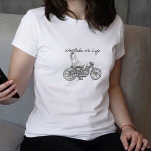 May include: White t-shirt featuring a black and white illustration of a person sitting on a motorcycle. The text "No Bike No Life" is above the motorcycle. The shirt is made of a soft material.