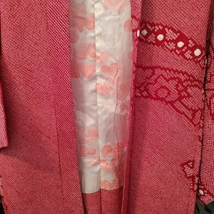 May include: A red kimono with a textured pattern and white accents. The interior lining features a floral design in shades of pink. The garment has long sleeves and a traditional Japanese design.