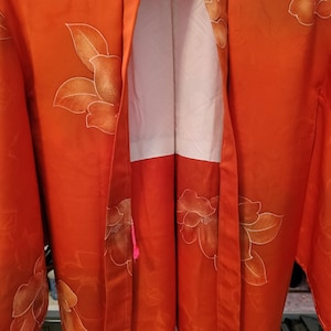 May include: An orange kimono with a floral pattern. The kimono features large, stylized flowers in shades of orange and white. The interior lining is white with a subtle geometric design. A pink tassel hangs from the garment.